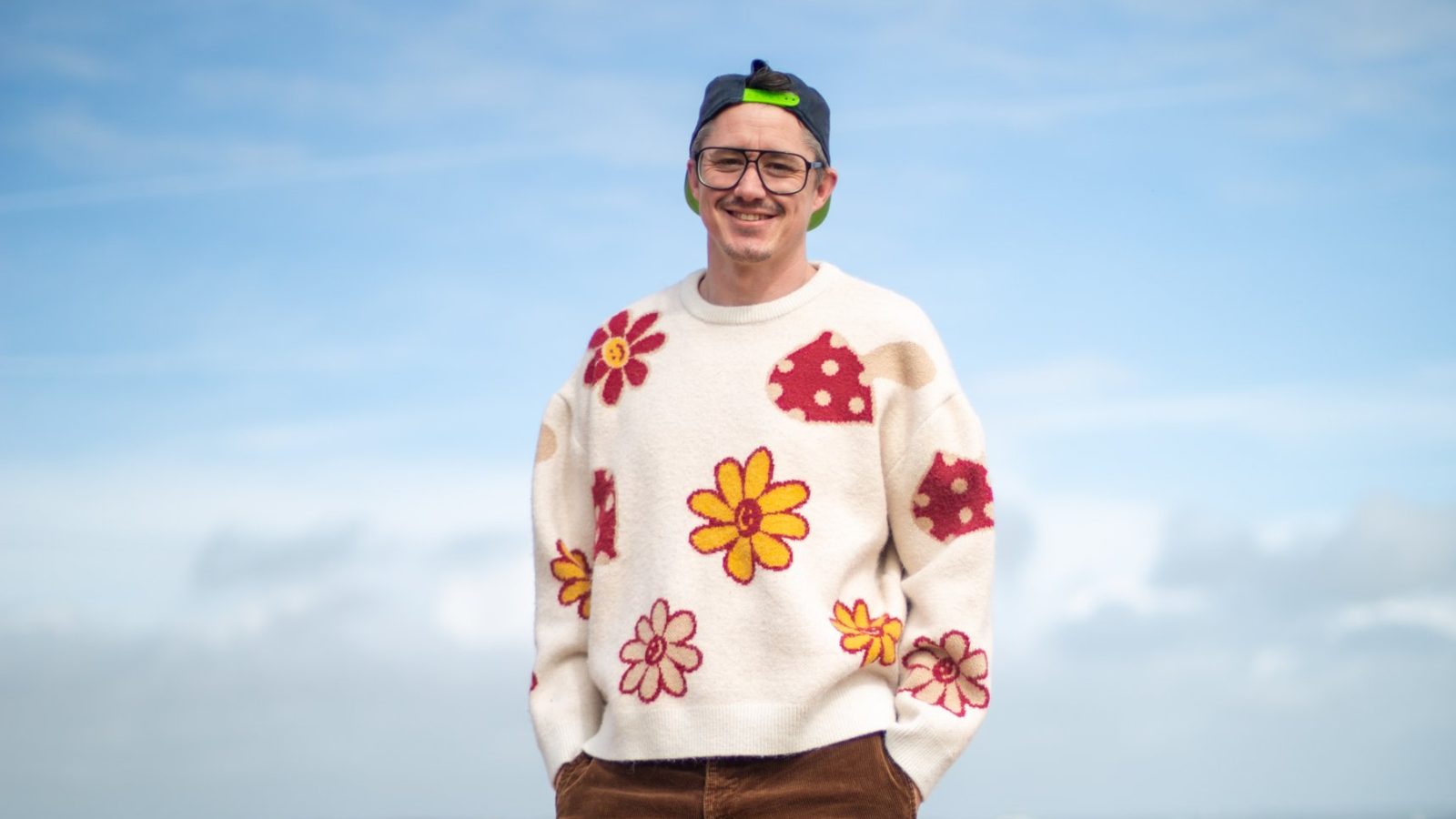 John McKie smiling on a shingle beach in a floral jumper and backwards cap